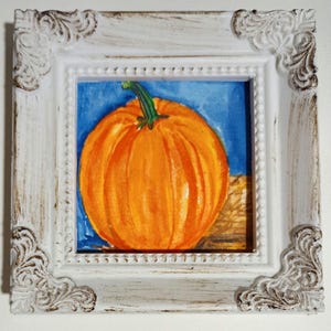 May include: A framed watercolor painting of a vibrant orange pumpkin with a green stem. The artwork is set against a blue background and a tan surface, enclosed in a white, ornate frame with decorative corners. The painting captures the essence of autumn.