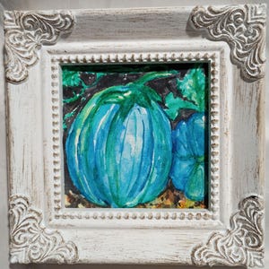 May include: A framed watercolor painting of two blue pumpkins with green leaves. The painting is in a white, ornate frame with decorative corner accents. The pumpkins are painted in shades of blue and teal.