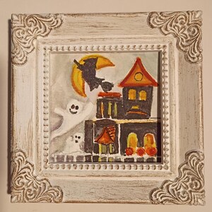 May include: A square painting in a distressed white ornate frame featuring a dark house with orange roofs, a yellow crescent moon, a black silhouette of a witch on a broomstick, and two white ghosts against a light gray background.