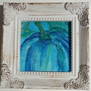 May include: A framed painting of a blue pumpkin with green leaves and stem. The artwork is in a white, ornate frame with decorative corners. The painting features shades of blue and green.
