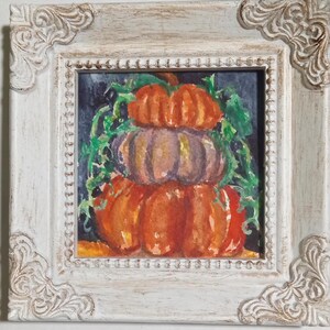 May include: A framed watercolor painting of three stacked pumpkins in shades of orange and purple, with green vines. The painting is set against a dark background and framed in a white, ornate, distressed-style frame. A fall-themed artwork.