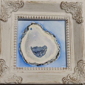 May include: A framed watercolor painting of an oyster shell. The shell is painted in shades of gray, blue, and white, set against a light blue background. The frame is white with ornate corner details.