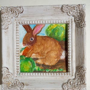 May include: A framed painting of a brown rabbit eating a carrot, set against a backdrop of green foliage and a blue sky. The artwork is enclosed in a white, ornate, distressed-style frame, adding a touch of rustic charm.