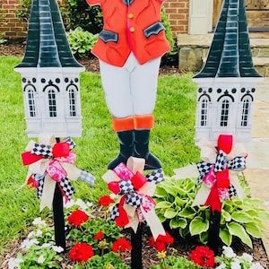 May include: Garden ornaments: a figure in a red coat and hat, flanked by two steeple-shaped posts. The posts are white with black roofs and decorative bows. Red and white flowers are planted in the garden bed.