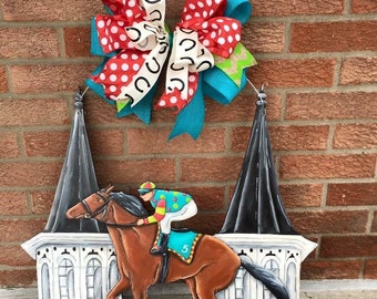 May include: A wooden door hanger with a painted image of a horse and jockey racing. The horse is brown and the jockey is wearing a colorful jersey. The door hanger is decorated with a large red and white polka dot bow with a turquoise ribbon.