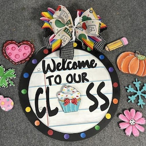 May include: Welcome to our class sign with a black and white polka dot border. The sign features a cupcake with blue and white frosting and sprinkles. The sign is surrounded by various decorations including a heart, a shamrock, a snowflake, a flower, a pencil, and a pumpkin.