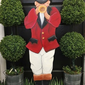 May include: A wooden cutout of a person in a red jacket, white pants, and black boots, blowing a horn. The figure is flanked by green topiary trees in metal buckets, with a black door in the background. The figure is wearing a black helmet.