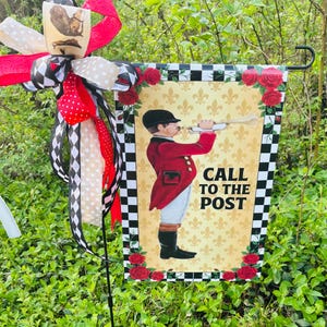 Puede incluir: Una bandera de jardín decorativa con un hombre con abrigo rojo tocando una trompeta, con el texto "CALL TO THE POST". La bandera tiene un borde a cuadros blanco y negro, rosas rojas y un lazo decorativo.
