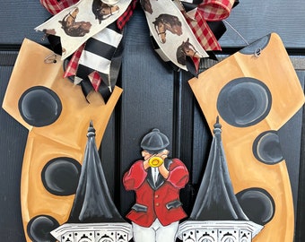 May include: A wooden horseshoe-shaped door hanger with a black and white checkered bow, a white bow with horse silhouettes, and a red and white checkered bow. The horseshoe is painted gold with black polka dots and features a black and white illustration of a person in a red jacket and white pants holding a trumpet. The horseshoe is flanked by two black and white buildings with white windows.