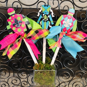 May include: Four colorful jockey silks with bows, in a clear vase with green moss. The silks are pink and green with a fleur-de-lis, blue and white with a fleur-de-lis, green and black with white polka dots, and blue and white with a bow tie.