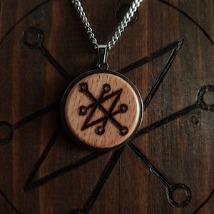 Könnte beinhalten: Ein runder Holzanhänger mit einem schwarzen geometrischen Design, eingefasst in einen silberfarbenen Metallrahmen, hängt an einer silbernen Kette. Der Anhänger wird vor einem dunklen Holzgrund mit geätzten Linien und Formen präsentiert.