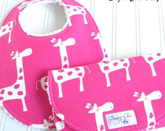 Pode incluir: Um conjunto rosa de babete e muda-fraldas para bebé com um padrão de girafas brancas. O babete tem um forro de tecido atoalhado branco e uma forma arredondada. O muda-fraldas é retangular e dobrável. O texto "Sugar Chic Baby" é visível.