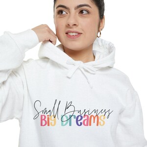 May include: White hoodie featuring the text "Small Business Big Dreams" in a script font and colourful block letters. The hoodie has a drawstring hood and long sleeves. The person in the image is wearing the hoodie.