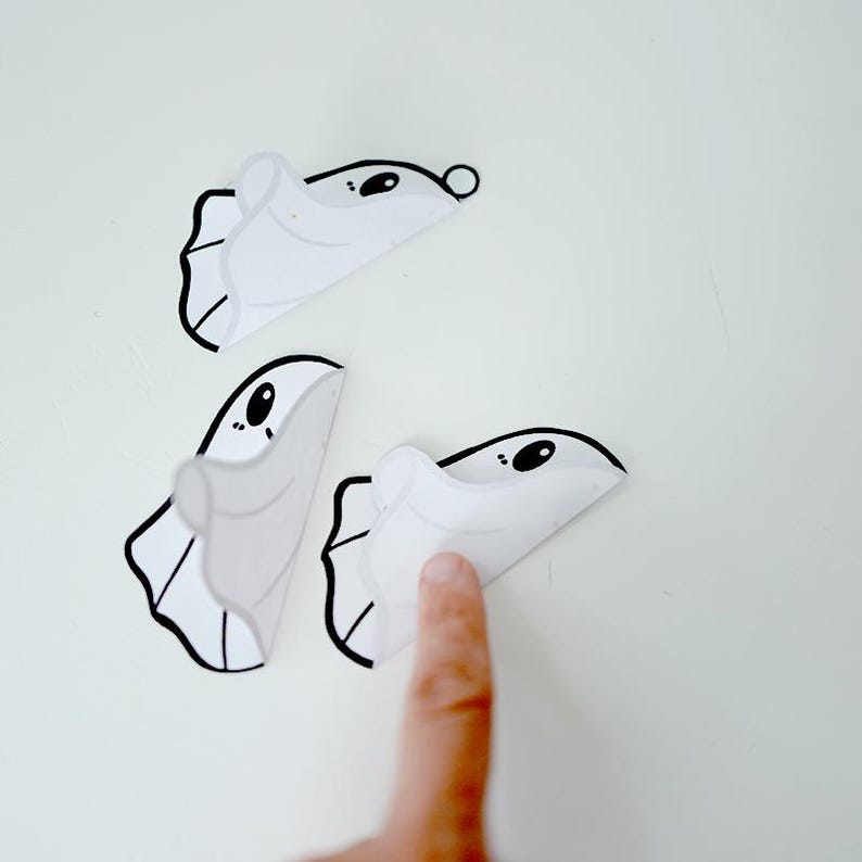K&ouml;nnte beinhalten: Drei wei&szlig;e Papier-Geister-Ausschnitte mit schwarzen Umrissen und Augen. Die Geister sind in der Mitte gefaltet und die Oberseite eines Geistes wird von einem Finger gehalten.