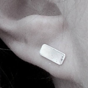 May include: A close-up, grayscale image of a rectangular silver earring. The earring is a small, flat rectangle, and is worn on the ear. The image also shows the ear and some hair.