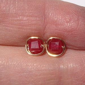 May include: A pair of gold-colored stud earrings, each featuring a square, deep red gemstone. The gemstones are set within a delicate gold wire frame, creating a classic and elegant design. The earrings are shown close-up.