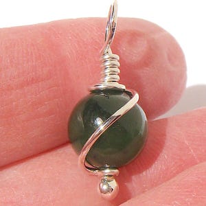 May include: A dark green, round gemstone pendant with a silver wire wrap and a silver loop. The pendant is held in a hand against a white background. The gemstone is approximately 0.5 inches in diameter.