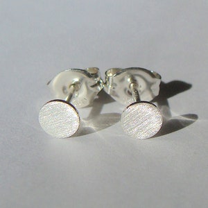 May include: A pair of silver stud earrings with a brushed finish. The earrings are round and have a simple design.