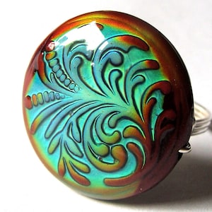 May include: A dichroic glass ring with a round, iridescent cabochon. The cabochon features a swirling, floral design in shades of teal, orange, and brown, set on a silver band. The glass reflects light, creating a vibrant, colorful effect.