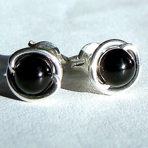 May include: A pair of stud earrings featuring a polished black gemstone set in a silver-toned, swirling design. The earrings have a classic, minimalist style, perfect for everyday wear. The gemstones are round and have a glossy finish.