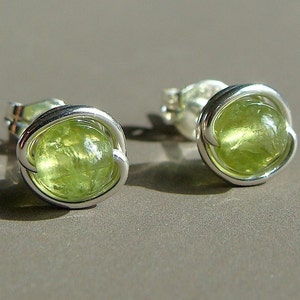 May include: A pair of silver earrings with green gemstone accents. The earrings are made with a simple wire design and feature a round, green gemstone in the center.