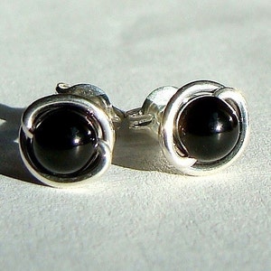 May include: A pair of stud earrings featuring a polished black gemstone set in a silver-colored, looped metal design. The earrings are round and have a simple, elegant style, suitable for everyday wear.