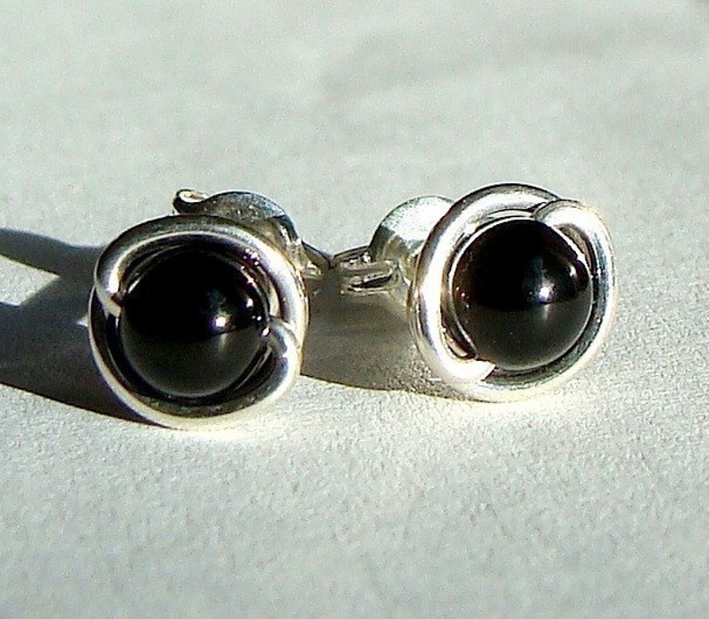 May include: A pair of stud earrings featuring a polished black gemstone set in a silver-colored, looped metal design. The earrings are round and have a simple, elegant style, suitable for everyday wear.