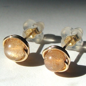 May include: A pair of gold earrings with a round, light brown sunstone gemstone set in a gold wire cage. The earrings have a simple, elegant design.
