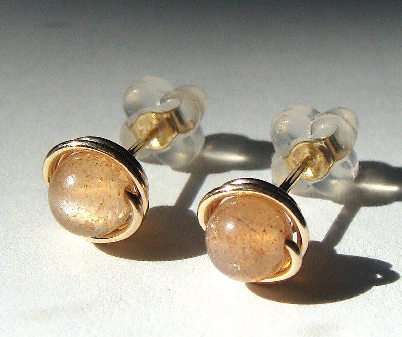May include: A pair of gold earrings with a round, light brown sunstone gemstone set in a gold wire cage. The earrings have a simple, elegant design.