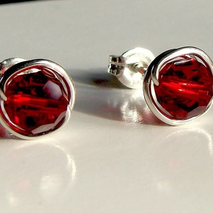 May include: A pair of stud earrings featuring round, faceted, deep red crystals. Each crystal is encased in a circular, polished silver-toned wire frame. The earrings include standard silver-toned post backs for secure attachment to the earlobe.