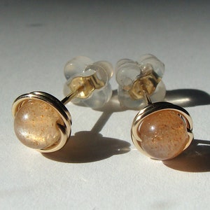 May include: A pair of gold sunstone earrings with a round, orange-brown sunstone set in a gold wire frame. The earrings have clear plastic backs.