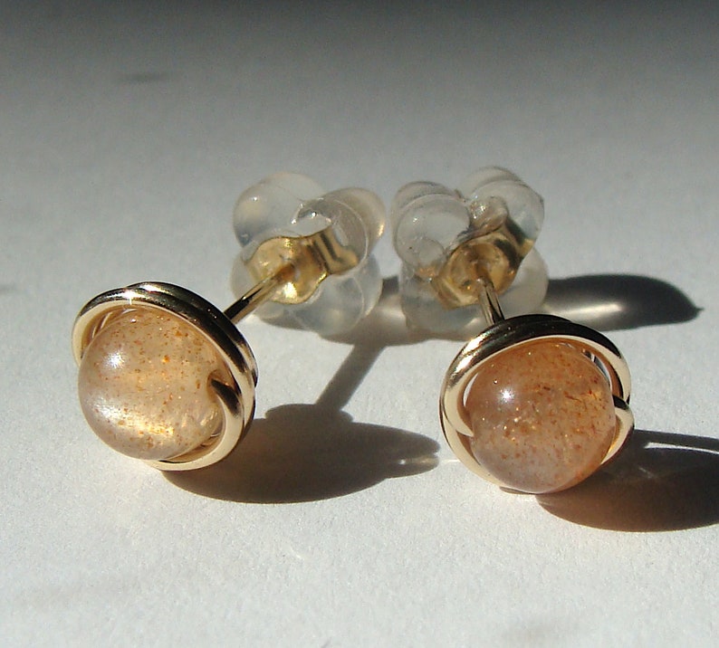 May include: A pair of gold sunstone earrings with a round, orange-brown sunstone set in a gold wire frame. The earrings have clear plastic backs.