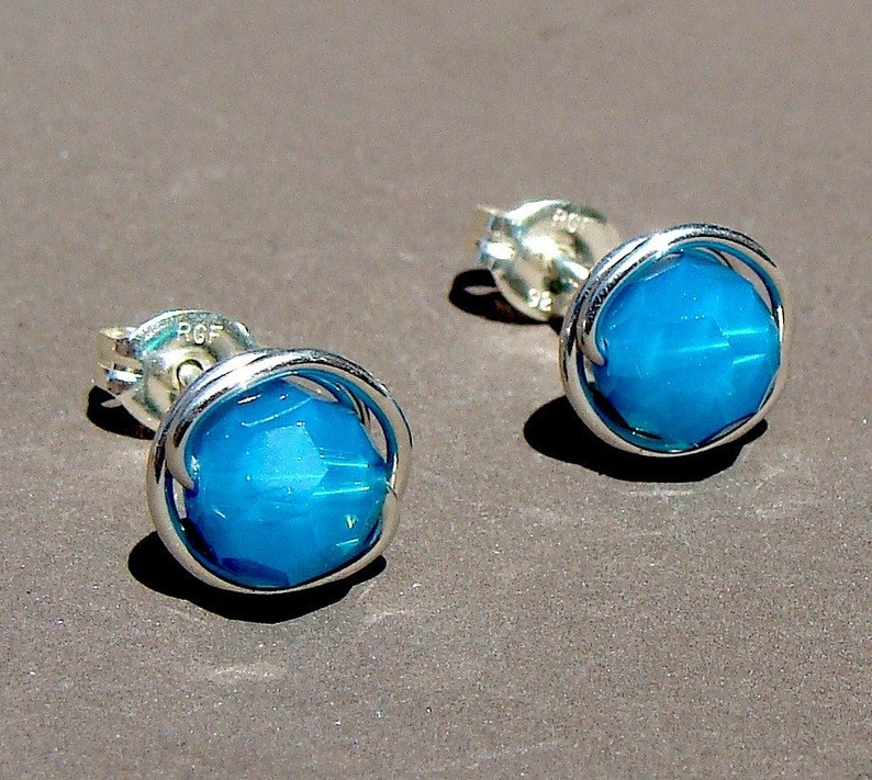 May include: A pair of silver stud earrings with a turquoise blue gemstone in each. The gemstones are surrounded by a silver wire frame.