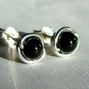 May include: A pair of stud earrings featuring a polished black gemstone set in a silver wire wrap. The earrings have a simple, elegant design, with a round black stone and a silver metal setting. The earrings are displayed on a light gray surface.