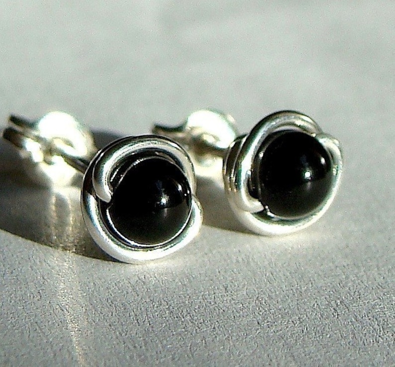 May include: A pair of stud earrings featuring a polished black gemstone set in a silver wire wrap. The earrings have a simple, elegant design, with a round black stone and a silver metal setting. The earrings are displayed on a light gray surface.