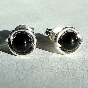 May include: A pair of silver stud earrings, each featuring a glossy black, round gemstone. The gemstones are set within a decorative, swirling silver frame. The earrings are displayed on a light gray surface.