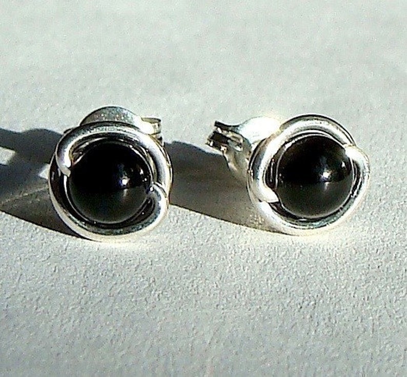 May include: A pair of silver stud earrings, each featuring a glossy black, round gemstone. The gemstones are set within a decorative, swirling silver frame. The earrings are displayed on a light gray surface.