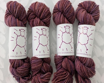 Puede incluir: Cuatro madejas de hilo teñido a mano en tonos burdeos, morados y marrones. Cada madeja está envuelta con una etiqueta blanca con una ilustración de oveja y la dirección del sitio web www.lavendersheep.com. El hilo está dispuesto sobre una superficie gris claro.