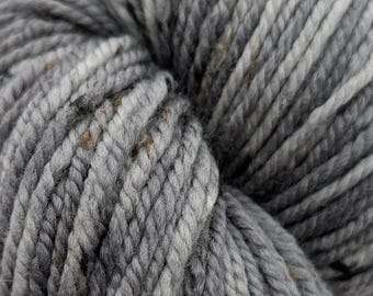 May include: Close-up of a skein of gray yarn with flecks of brown and white. The yarn is thick and textured, ideal for knitting or crocheting projects. The color is a mix of light and dark grays.