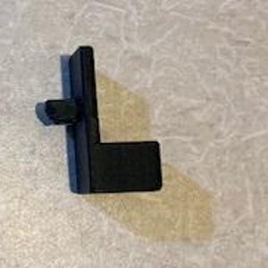 May include: A black, L-shaped plastic component with a small, rectangular projection. The piece is designed with a right angle and a small, square peg extending from one side. The item is likely a part of a larger mechanism or structure.
