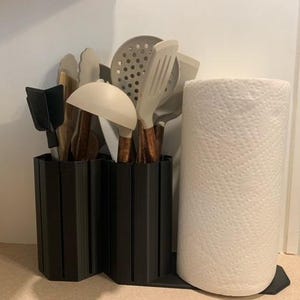 May include: A black kitchen utensil holder with a paper towel holder. The utensil holder contains various cooking tools with wooden handles and light-colored heads. A roll of white paper towels sits next to the utensil holder.
