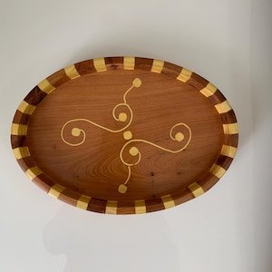 Peut inclure: Plateau de service ovale en bois avec un centre brun clair et une bordure décorative en bois brun clair et foncé alterné. Le plateau présente un motif tourbillonnant de couleur or au centre.