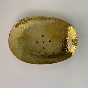 Peut inclure: Porte-savon ovale en laiton martelé. Le plat est de couleur dorée brillante et comporte cinq petits trous de drainage. Le porte-savon est conçu pour contenir un pain de savon. Dimensions en centimètres.
