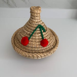 May include: A woven basket with a conical lid. The basket is made of natural-coloured straw and features a green stem with two red pom-pom cherries on the lid. The basket is likely used for storage or decoration.