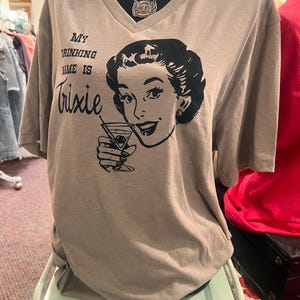 May include: A taupe V-neck t-shirt with the text "My Drinking Name is Trixie" and a black and white illustration of a woman holding a martini. The shirt is displayed on a mannequin.