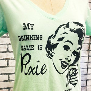 May include: A light green t-shirt with a black graphic print of a woman holding a glass. The text on the shirt reads "My Drinking Name Is Pixie".
