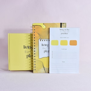 Può includere: Un set di tre quaderni gialli e bianchi con il testo "living on the FATTER SIDE priorities" e "living but playing" sulle copertine. I quaderni sono su una superficie rosa.