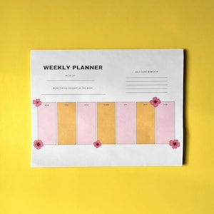 May include: A white weekly planner with the text "WEEKLY PLANNER" and "SELF CARE MOMENTS" on a yellow background. The planner has pink and gold sections for each day of the week, decorated with pink flower illustrations.