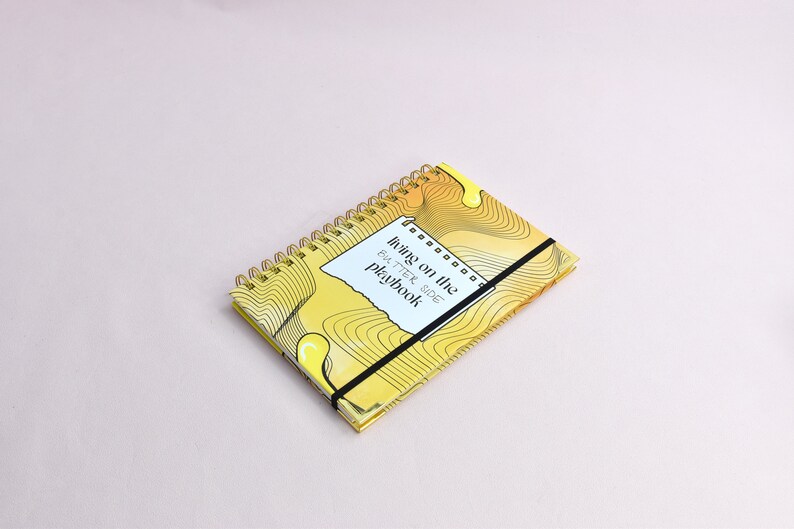 Yellow, guided journal lying flat on a surface, enhanced with gold binding, gold corners, elastic holder.