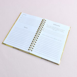 Guided journal displays two pages of content inside of the journal. Features a reflection page and a page with a date and notes section; gold binding.